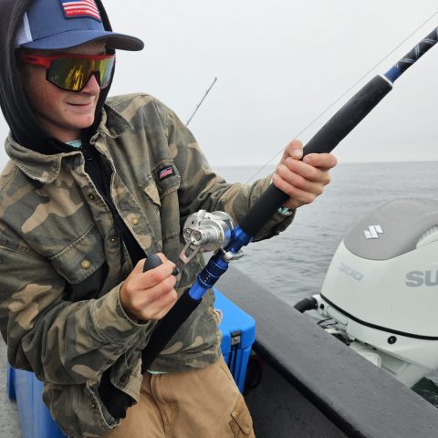 Person in camo jacket fishing on a boat with a blue fishing rod.