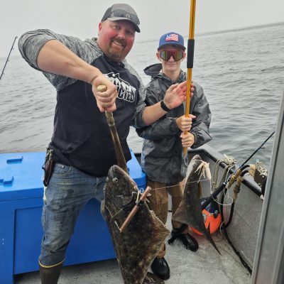 Two people on a boat holding fishing poles with large fish caught over the water.