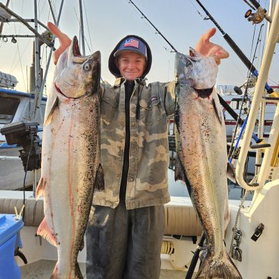 Person on a boat holding two large fish, with fishing rods in the background.