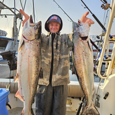 Person on a boat holding two large fish, with fishing rods in the background.