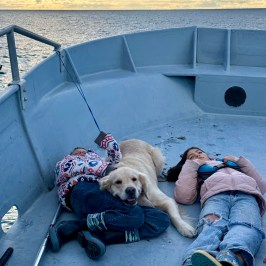 Two kids and a dog resting on a boat deck with the ocean in the background.