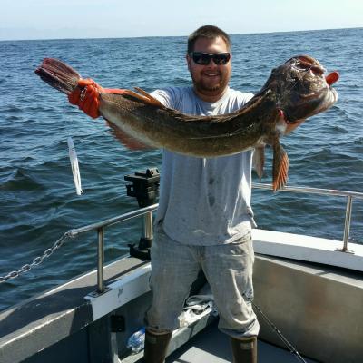 Person wearing sunglasses holding a large fish on a boat with ocean in the background.