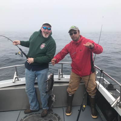 Two men on a boat holding fishing rods, one with a fish, on a foggy day at sea.