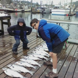 Two children on a dock posing with fish lined up on the wooden boards, boats in the background.