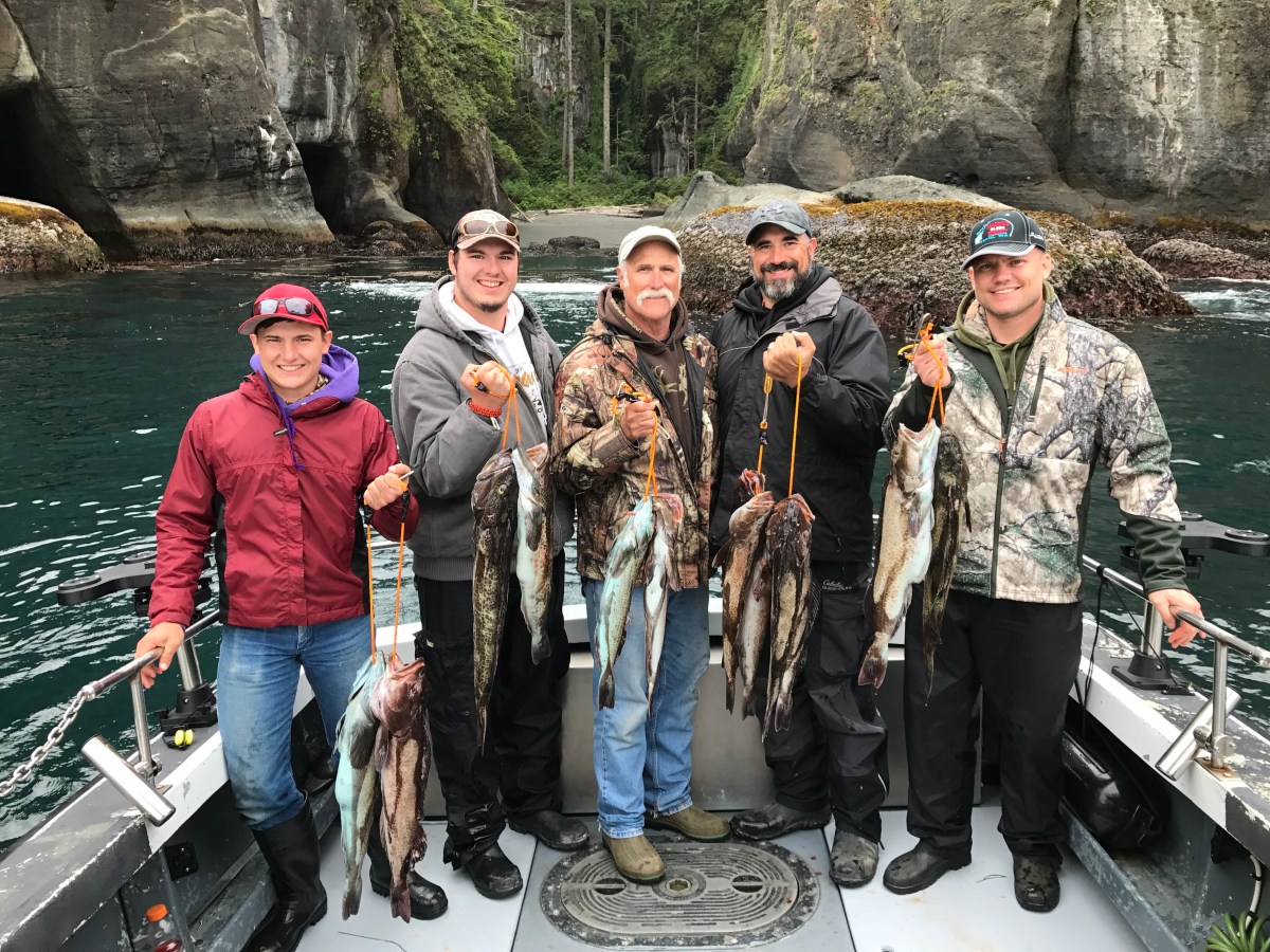 Five people on a boat holding fish with rocky cliffs and trees in the background.