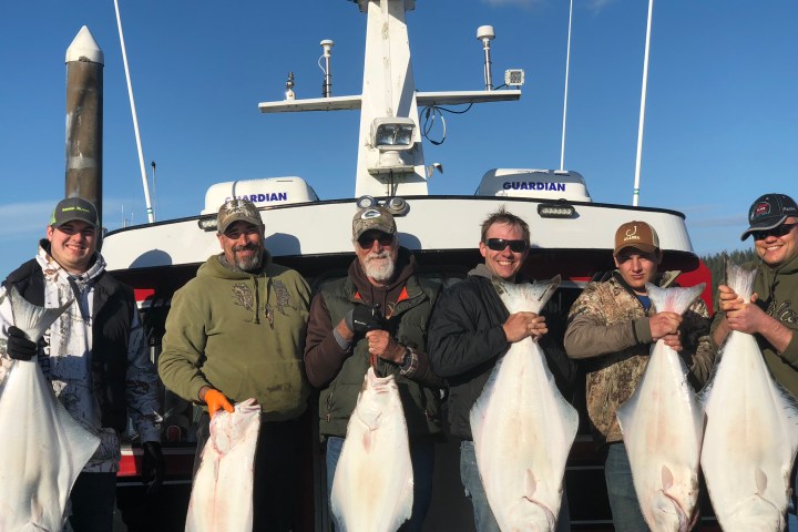 Group of six men holding large fish in front of a boat named Guardian.