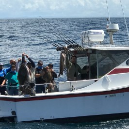 People on a boat holding up large fish with fishing rods visible and ocean in background.