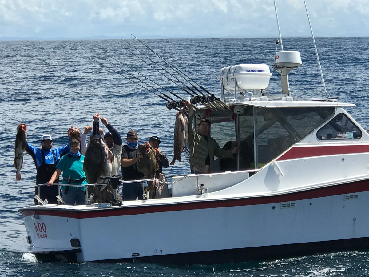 People on a boat holding up large fish with fishing rods visible and ocean in background.