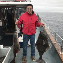 Person in red jacket holds two large fish on a boat deck with ocean in the background.
