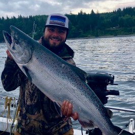 Man in camouflage holding a large fish on a boat with a lake and trees in the background.