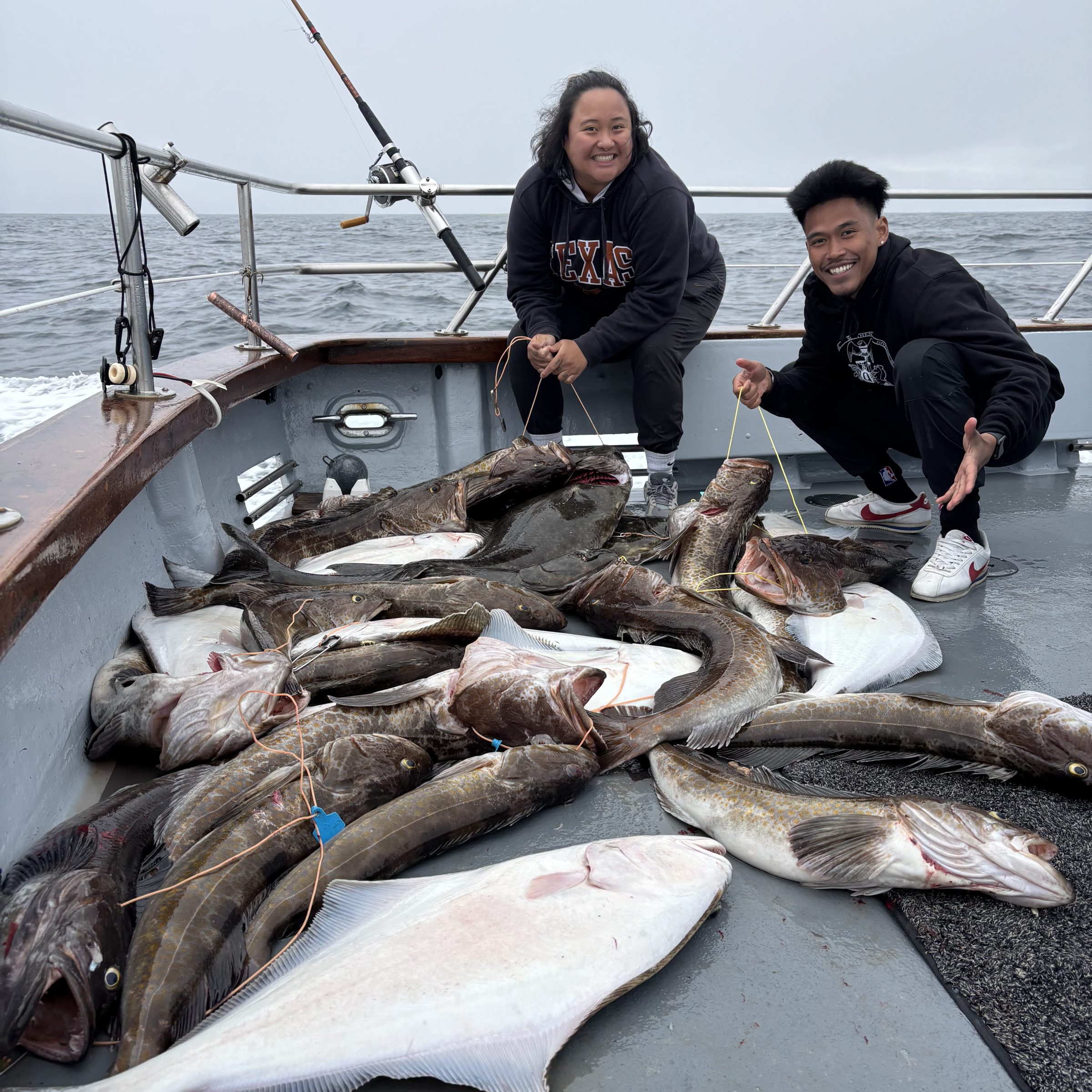 Two people on a boat deck with a large catch of fish, including halibut, on a cloudy day.