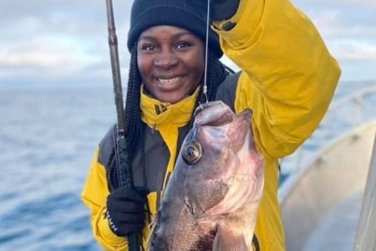 Person in yellow jacket holding a large fish on a boat with the sea in the background.
