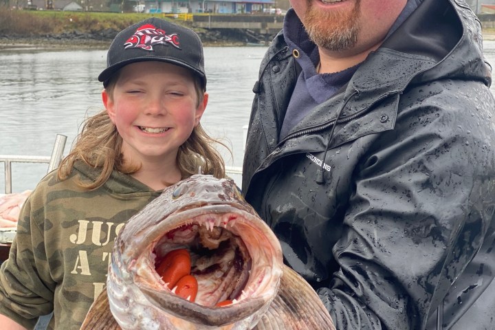 Two people smiling, holding a large fish with an open mouth on a rainy day near a body of water.