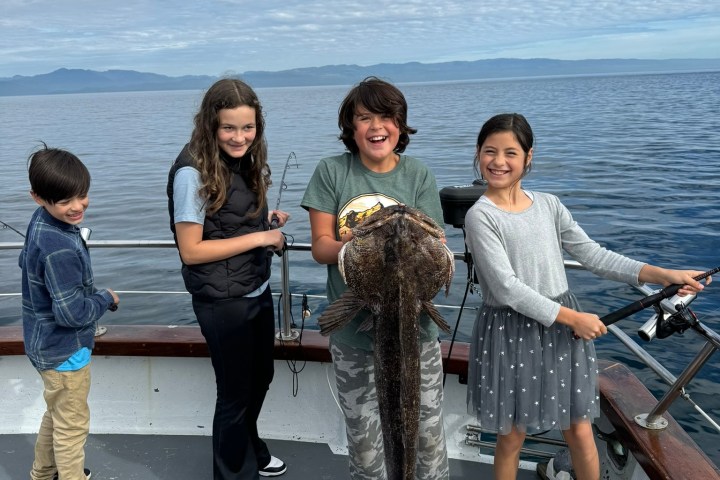 Four smiling children on a boat; one holds a large fish, cloudy sky and ocean in background.