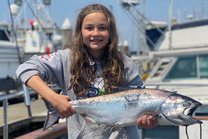 Person holding a large fish on a boat dock with fishing boats in the background.